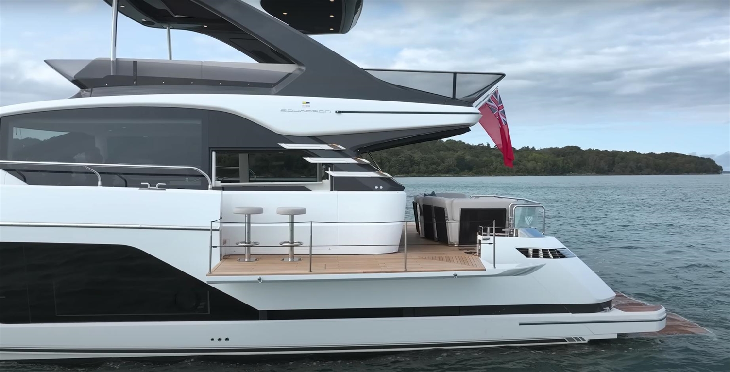 Modern yacht with sleek design, featuring outdoor seating, bar stools, and British flag on the rear in a serene water setting.